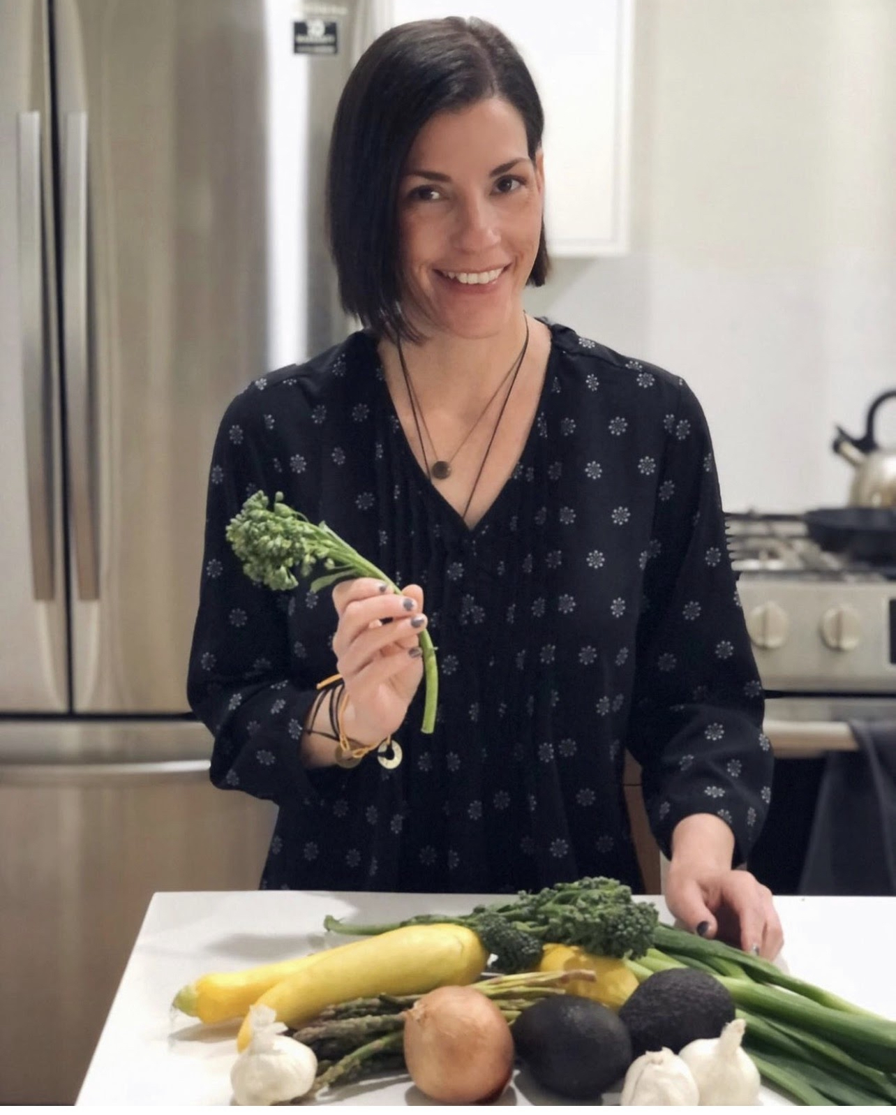 Jen Yundt Coles, The SIBO Coach Jen Yundt Coles in her kitchen holding fresh broccoli surrounded by colorful vegetables.